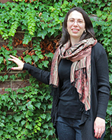 Image of Jennifer Gipson outdoors in front of an ivy-covered walls wearing a black shirt and colorful scarf. Her hand reaches out to touch the ivy.