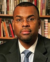 Portrait image of Eddie Glaude seated in front of book shelves wearing glasses, a black suit, and a blue tie