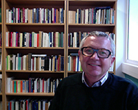 Portrait image of Simon Gaunt seated in front of bookshelves wearing glasses and a black sweater