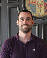 Portrait image of Louis Betty standing in front of a wooden wall and crest wearing a purple polo shirt