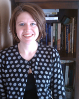 Portrait image of Caroline Boswell wearing a dotted black jacket standing in front of a book shelf