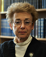 Portrait image of Rachel Feldhay Brenner wearing a white turtleneck, black sweater, and pin seated in front of blue books