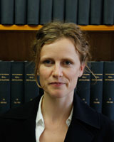Portrait image of Julie Gibbings wearing a white shirt and black jacket standing in front of book shelves lined with blue books