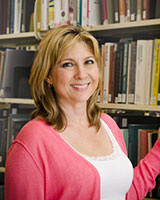 Portrait image of Marguerite Helmers standing amidst library book shelves wearing a white shirt and pink sweater