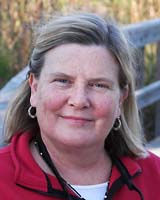 Portrait image of Michele Hilmes outdoors wearing a red shirt and small hoop earrings standing in front of a wood fence.