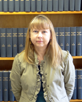Portrait image of Theresa Kaminski wearing a khaki jacket standing in front of book shelves