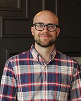 Portrait image of Adam Mandelman wearing a flannel shirt and black glasses
