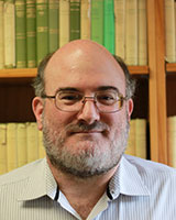 Portrait image of Gregory Milton standing in front of book shelves wearing glasses and a buttoned shirt
