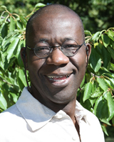 Portrait image of Tejumola Olaniyan outdoors in front of green plants wearing a white shirt and glasses.