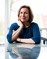 Portrait image of Mary Louise Roberts seated at a glass table with her arms crossed and hand at neck wearing a blue shirt