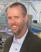 Portrait image of James H. Sweet outdoors in front of a dock wearing a striped shirt and brown jacket