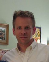 Portrait image of Colin Wilder wearing a white shirt standing in front of hung art works