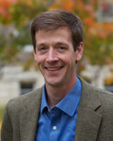 Portrait image of Lee Willis outdoors with fall leaves behind him wearing a blue shirt and brown jacket