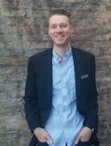 Portrait image of Victor Lenthe standing in front of a brick wall wearing a blue shirt and dark jacket