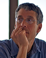 Portrait image of Steven Nadler wearing glasses and a blue shirt with his hand on his chin in front of a window