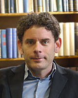 Portrait image of Brian Sandberg wearing a dark jacket seated in front of book shelves
