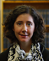 Portrait image of Stephanie Spadaro in front of book shelves with shoulder-length curly hair wearing a black shirt and white-and-black scarf