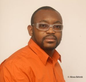 Portrait image of Jean-Pierre Bekolo wearing an orange shirt and clear glasses