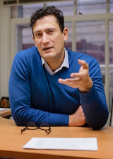 Portrait image of Pablo Gomez seated at a table speaking, wearing a blue shirt and gesturing with his left hand