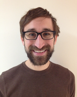 Portrait image of Andrew Zolides wearing a brown shirt and glasses standing in front of a white wall
