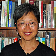 Portrait image of Christine Yano standing in front of book shelves wearing a black shirt and glasses