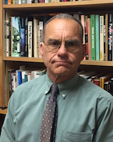 Portrait photograph image of Larry Nesper standing in front of book shelves wearing a green shirt and burgundy tie.