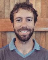 Portrait image of Nicholas Jacobson wearing a colored shirt standing in front of a wood-paneled wall