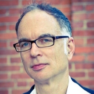 Portrait image of Ken Wissoker standing in front of a brick wall wearing a suit and dark-rimmed glasses