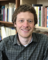 Portrait image of John Boonstra seated in front of book shelves wearing a buttoned shirt