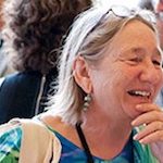 portrait image of Susan Friedman seated at an event table laughing with her hand at her chin. She wears a brightly colored shirt and large earrings and has her glasses pushed up on her head.