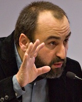 Portrait image of Costica Bradatan wearing a grey and black suit and speaking into a microphone with his hand held up