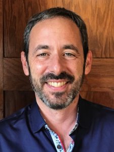 Portrait image of Stephen Kantrowitz wearing a blue shirt standing in front of a wood wall