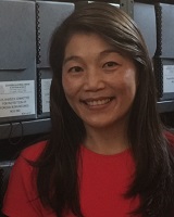 Portrait image of Cindy I-Fen Cheng wearing a red shirt standing in front of shelves of grey archival boxes