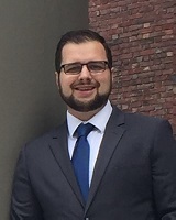 Portrait image of Daniel Hummel wearing a grey suit and blue tie standing in front of a brick wall