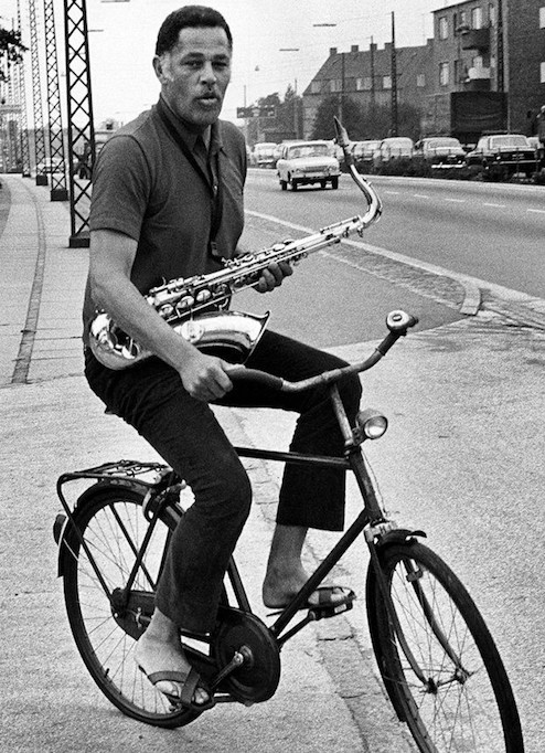 Image of Dexter Gordon on a bicycle carrying his saxaphone via a neck strap and one hand