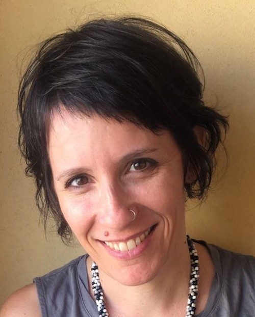 Portrait image of Keren Omry in front of a beige-yellow wall wearing a grey shirt and beaded necklace