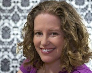 Portrait image of Rachelle Winkle-Wagner wearing a purple shirt in front of a black and white decorated wall.