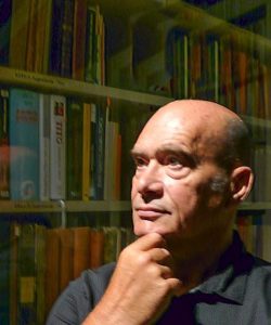 Portrait image of Toma Longinovic holding his chin standing in front of book shelves with a glass reflection in front of the shelves.