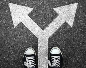 Image of two identical arrows pointing in different directions painted in white on black asphalt and two shoes a the base of the arrows.