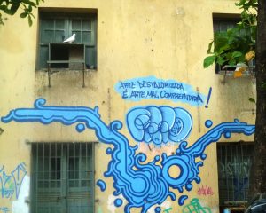 A photograph depicting graffiti on a residential building. An abstract, light blue, undulating shape lies below a spray painted message which reads, in Portuguese: "Un-valorized art is misunderstood art."