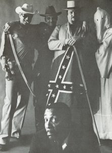 Black and white satirical image of Dick Gregory (a frowning Gregory appears in the bottom foreground of the image while a smiling group of men appear standing in the upper background, including a police officer, a hooded Klansman, a confederate civil war soldier holding a bayonet, and a man holding a confederate flag).