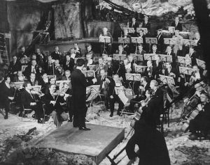 Black and White photo of Sergiu Celibidache conducting Beethoven’s Egmont overture in the ruins of the Philharmonie concert hall, musicians are wearing tuxedos and rubble is visible near conductor's podium