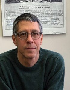 Portrait image of Steven Nadler wearing a dark green sweater seated in front of a poster