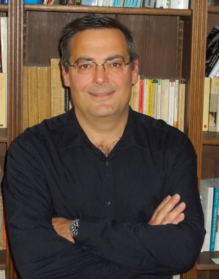 Portrait image of Paul-Alexis Mellet in standing in front of book shelves with his arms folded.