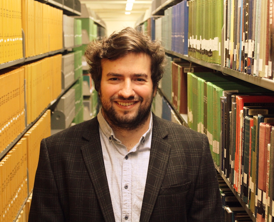 Portrait photo of Ari Sekeryan in a library aisle surrounded by book shelves.