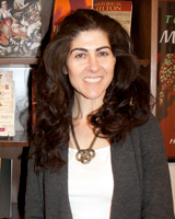 Portrait image of Stacy S. Klein standing in front of book shelves wearing a white shirt, grey sweater, and large necklace