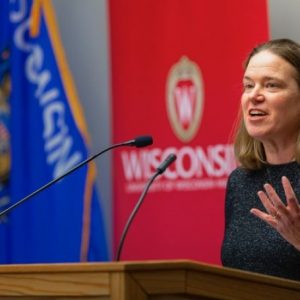 Image of Anna Andrzejewski speaking at a podium with two microphones in front of a Wisconsin state flag and a University of Wisconsin banner. She is wearing a grey shirt and gestures with her left hand