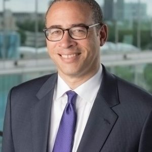 Portrait image of Jonathan Holloway wearing a grey suit, glasses, and purple tie standing in front of a mirror