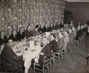 Black and White image of a long table with many men in suits seated around the table, looking at the camera
