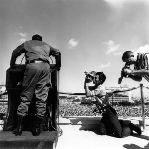 black and white image of the back of Fidel Castro speaking at a podium with men holding news cameras kneeling at the right side of the image